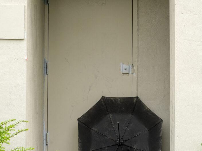 A black umbrella rests in a beige doorway with green foliage in the foreground.